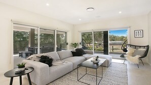 Ein modernes Wohnzimmer mit grauem Sofa, einem Couchtisch und einem schwarzen Sessel. Große Fenster bieten einen Blick ins Freie.
