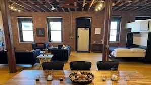 A spacious room with a wooden dining table, black chairs, and a large bowl of snacks. There are two windows with curtains, a door, and a bed with a white headboard. - Brick City Loft (Newark)