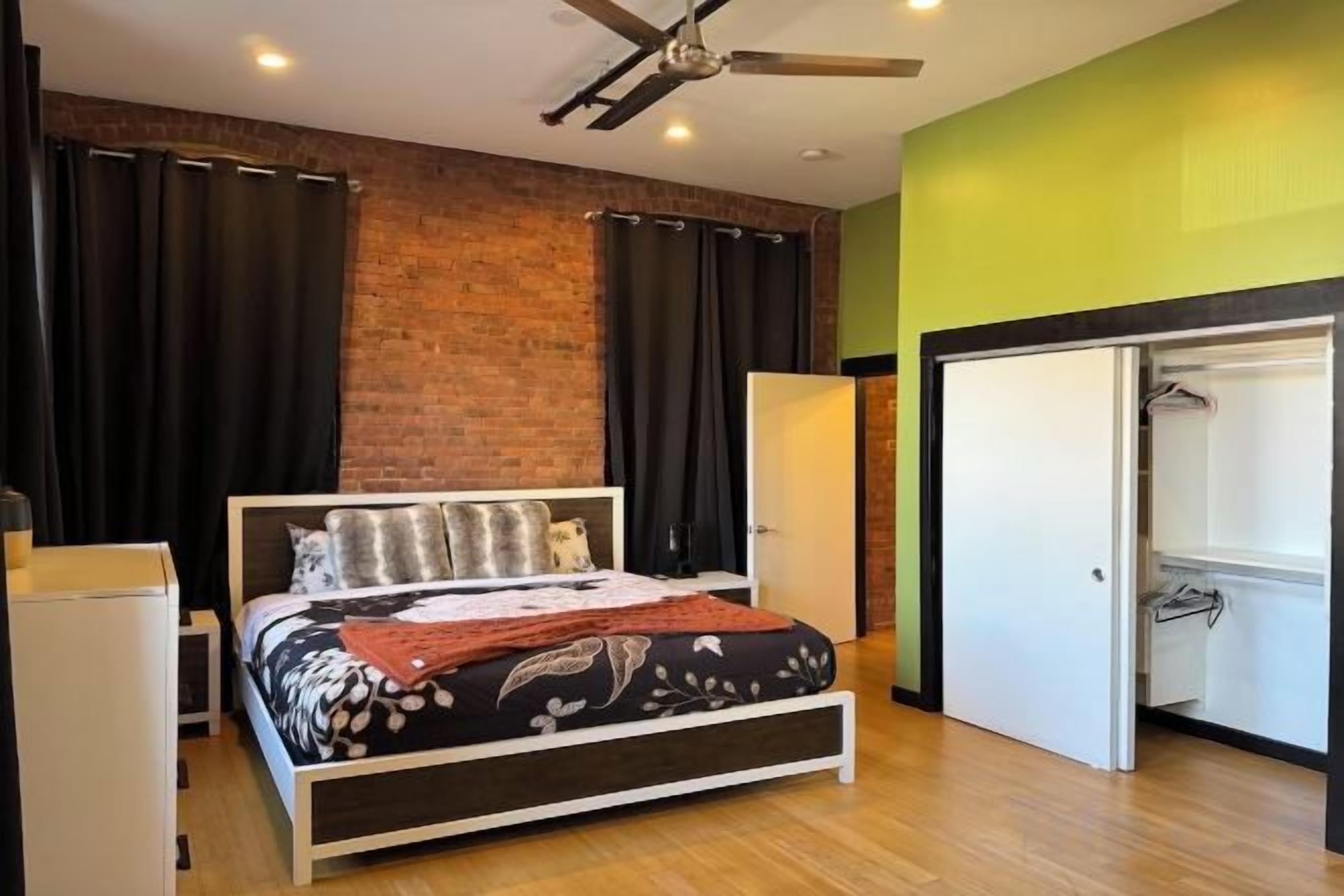 A bedroom with a bed, a wardrobe, a ceiling fan, and a brick wall.