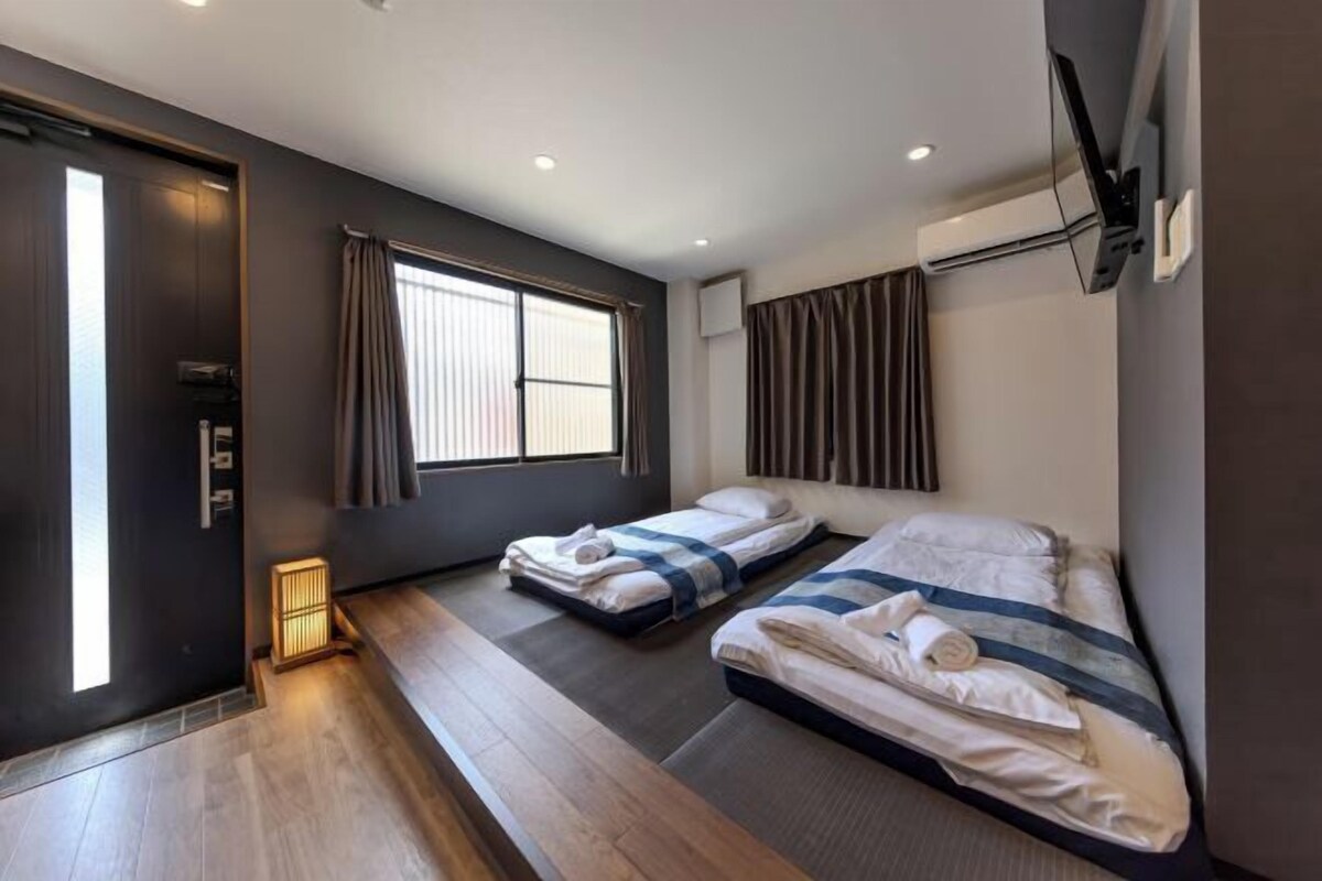 A hotel room with two beds, a wooden floor, a window with curtains, and a wall-mounted TV.