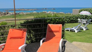 Ein Balkon mit zwei orangefarbenen Lounge-Stühlen und einem Tisch, der Blick auf eine felsige Küste und das Meer bietet.