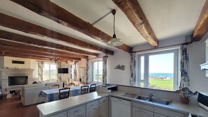 Eine Küche mit weißen Schränken, einem Kamin aus Naturstein, einem Essbereich mit Tisch und Stühlen sowie Meerblick durch große Fenster.
