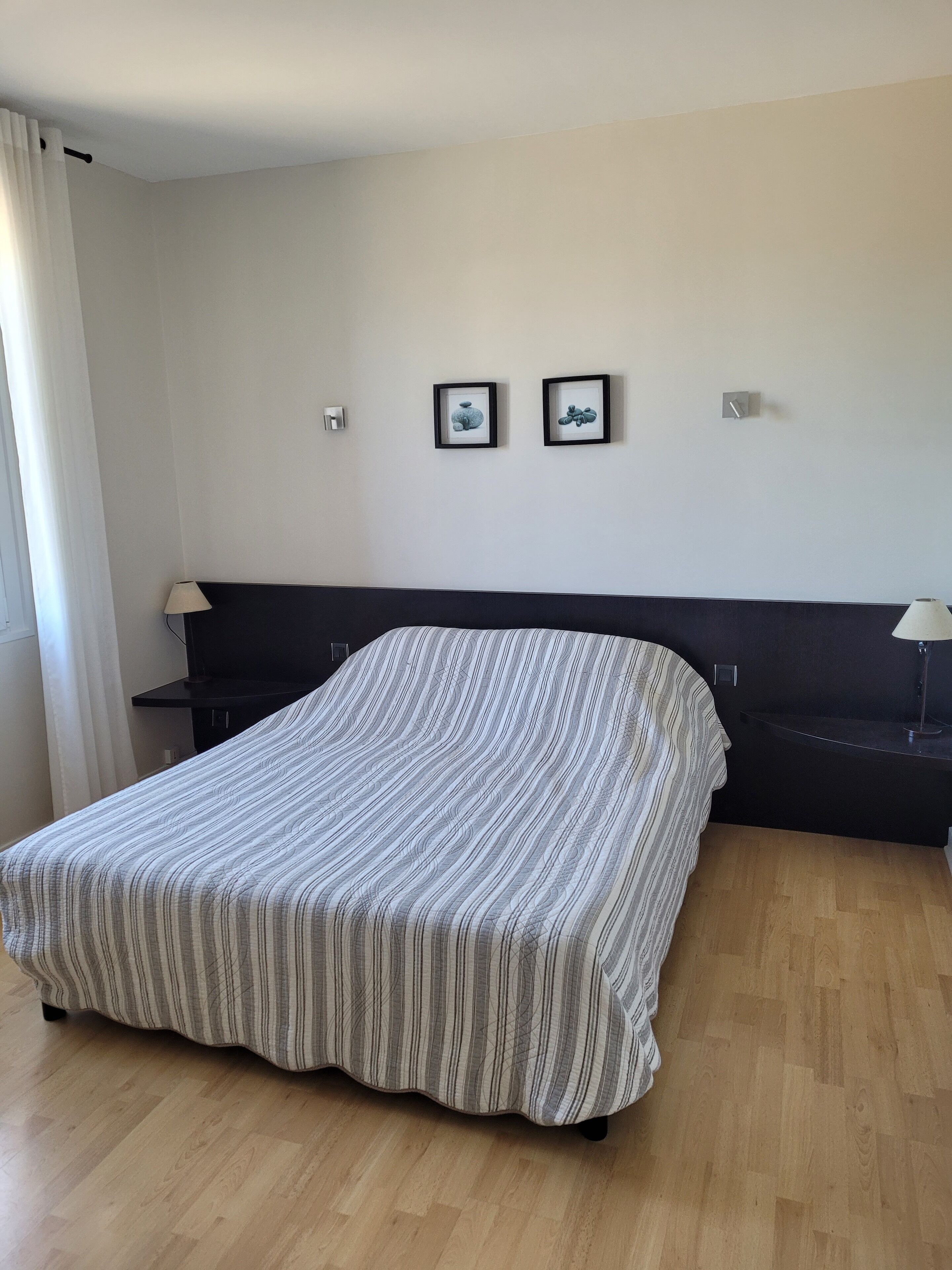 Une chambre à coucher comprenant un lit, deux tables de chevet, une commode, une horloge murale et deux tableaux encadrés.