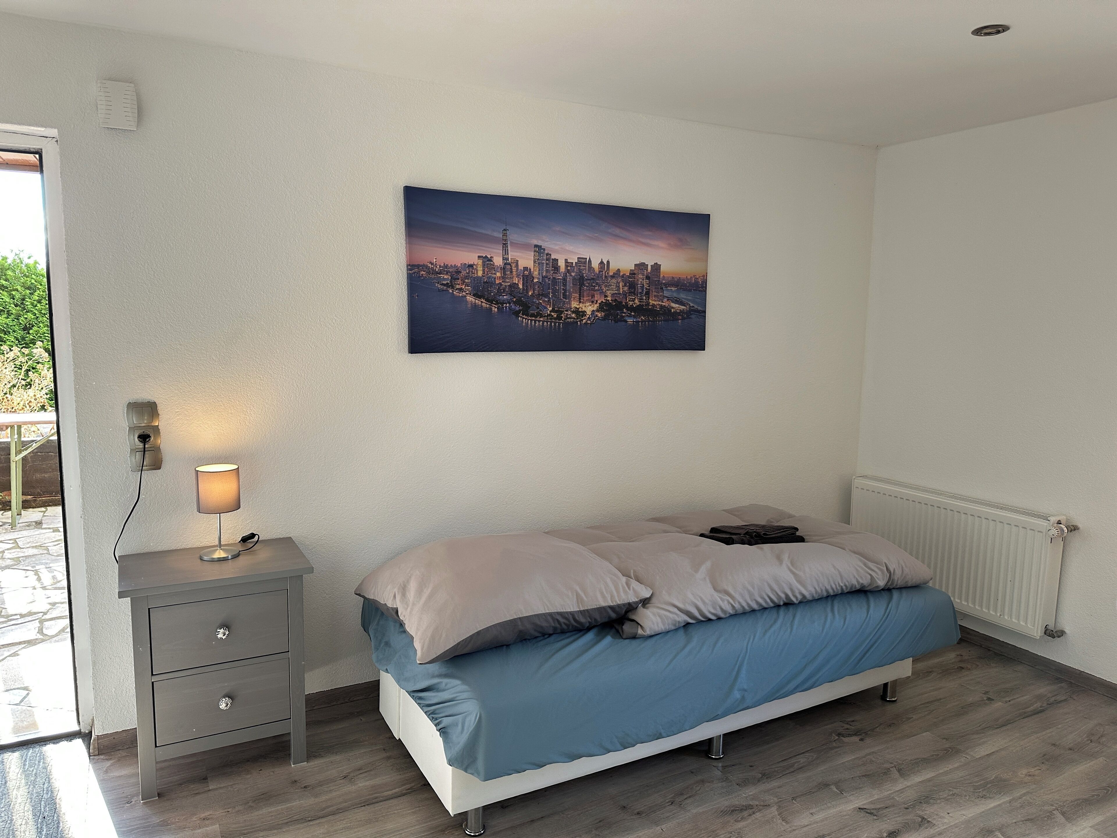 Un dormitorio con cama, mesita de noche, lámpara, radiador y un cuadro de una vista urbana.