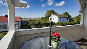 Uma varanda com mesa de vidro, uma planta em vaso com flores vermelhas e vista para uma casa branca e paisagem verde.