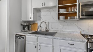 A kitchen with white cabinets, a marble backsplash, a stainless steel sink, a coffee maker, and a wooden cutting board. - King Bed 2BR Retreat, Private Garage (Kansas City)