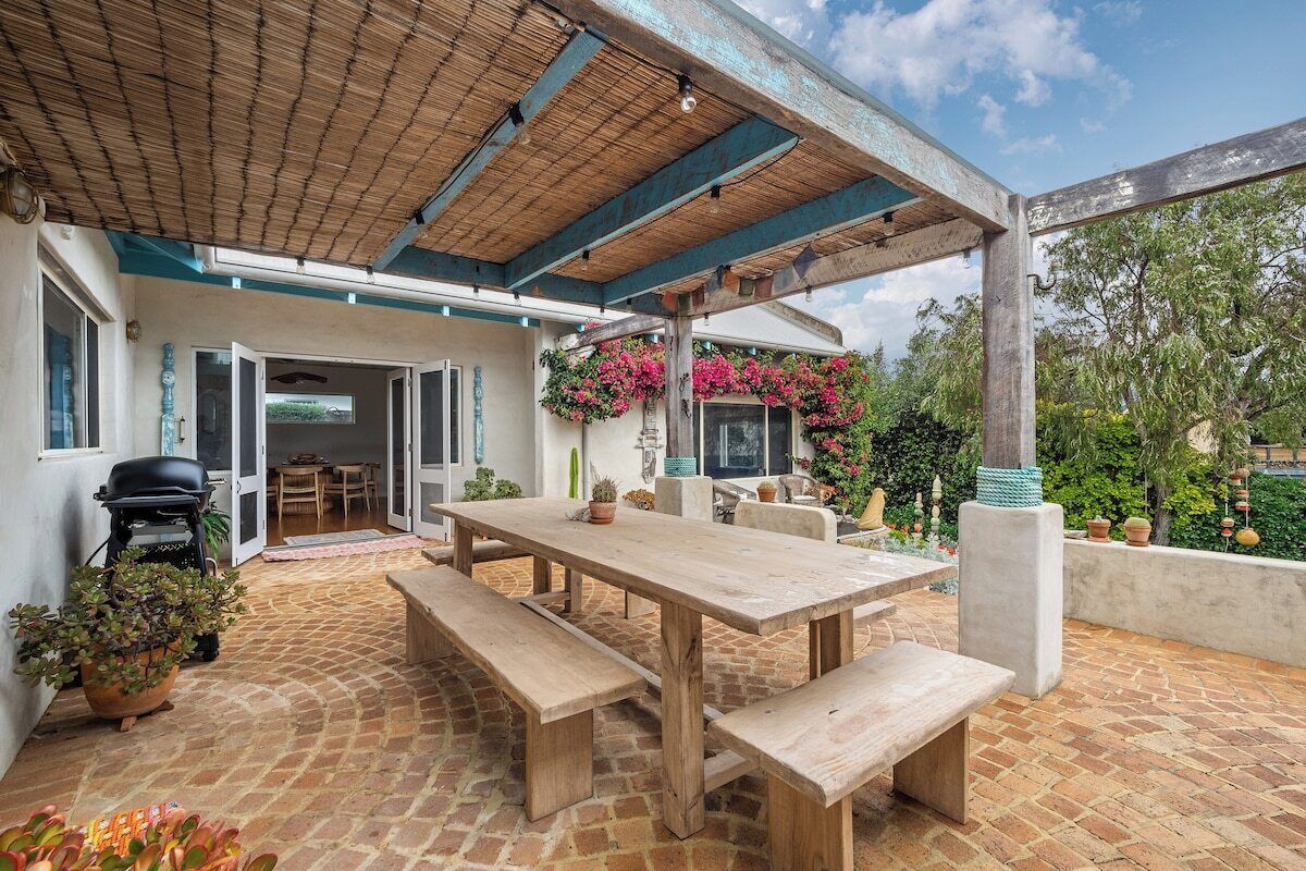 An outdoor patio with a wooden table and benches, surrounded by potted plants and a barbecue grill.