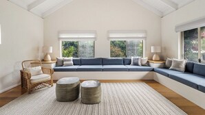 A room with a high ceiling, a built-in bench with cushions, two windows with blinds, a wicker chair, and two round ottomans. - Saltwood: Yallingup Garden Sanctuary by the Sea (Yallingup)