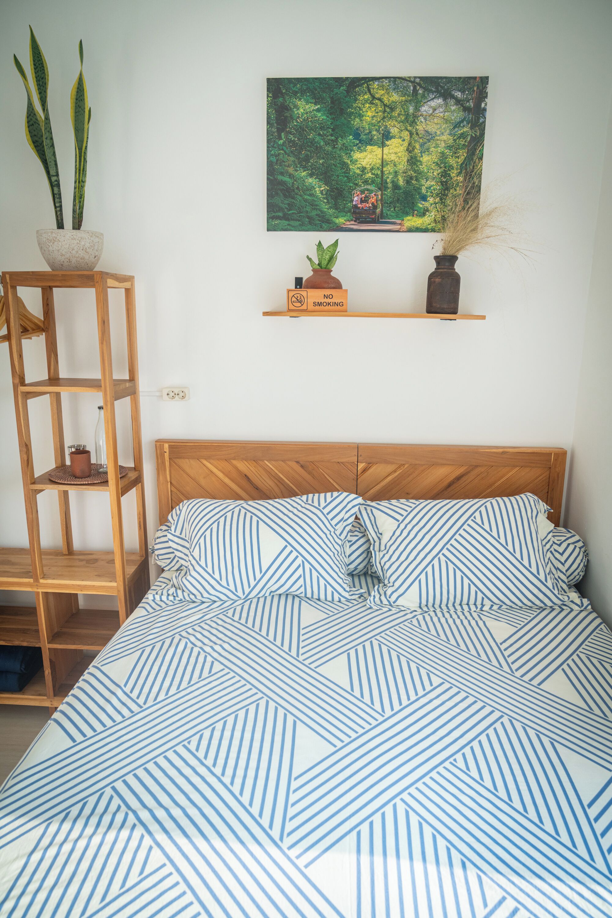 A bedroom with a wooden headboard, a bed with blue and white striped bedding, a wooden shelf with a potted plant, and a wall-mounted painting of a forest scene.