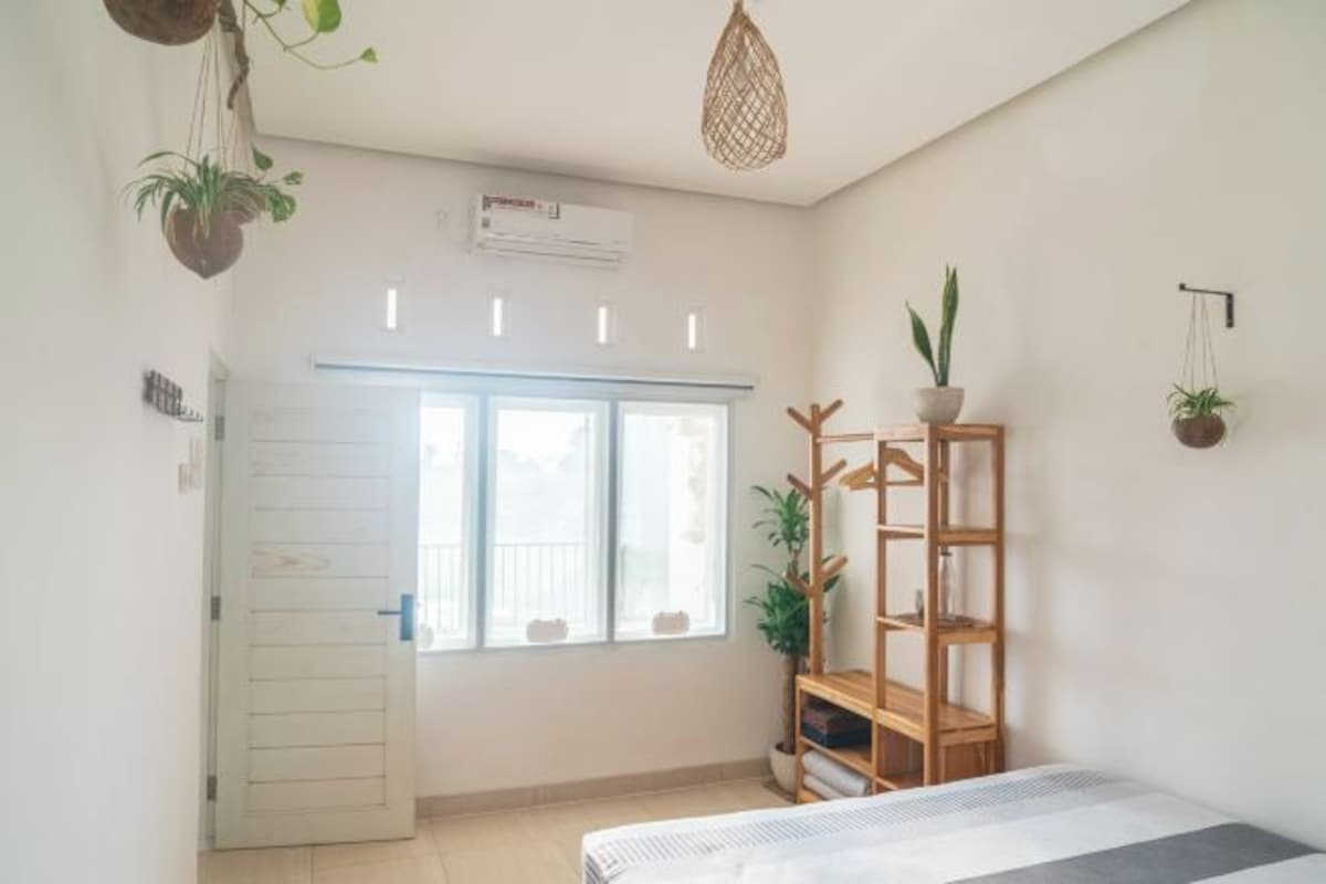 A room with a bed, a window, a wooden shelf, and hanging plants.