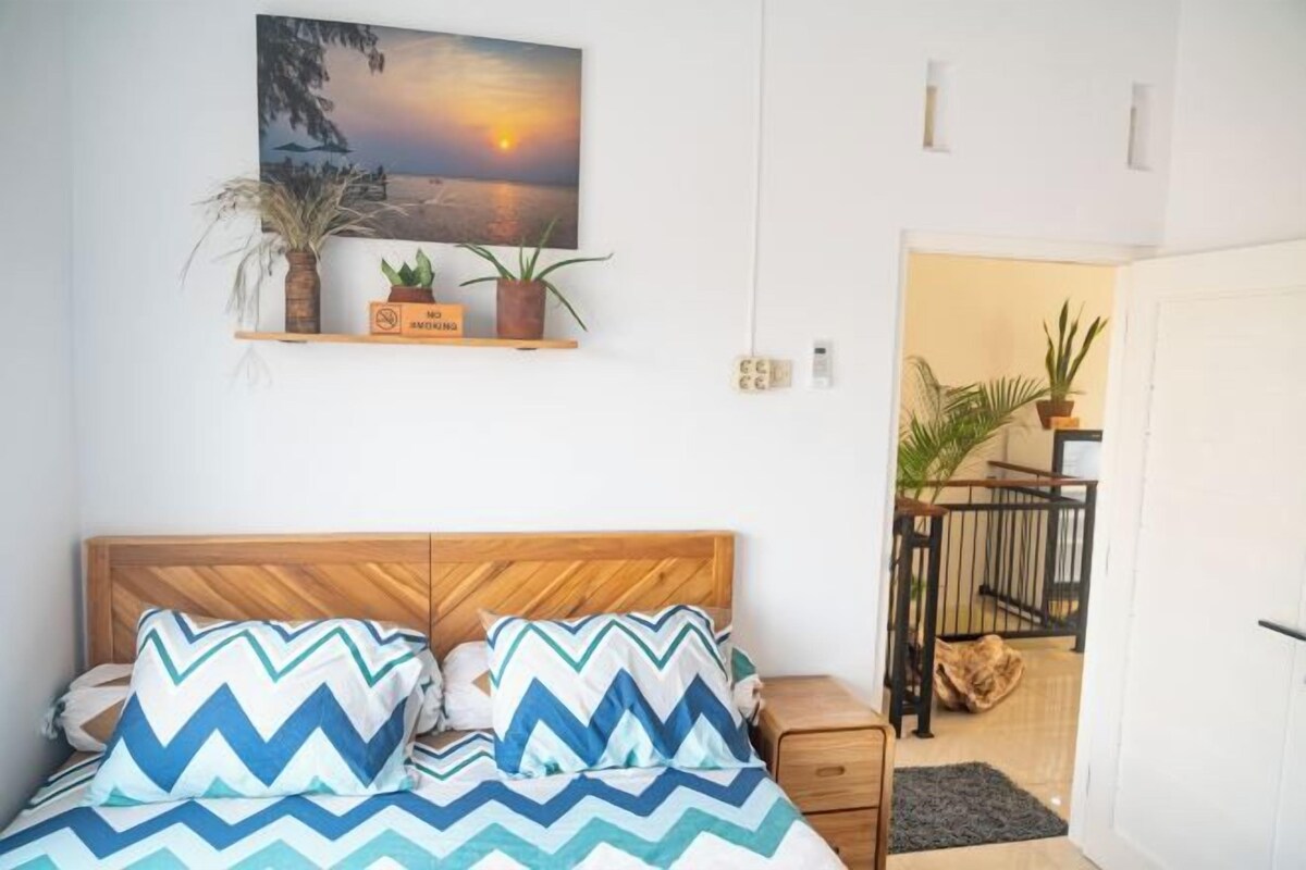 A bedroom with a wooden headboard, a bed with patterned bedding, a nightstand, a wall-mounted shelf with plants, and a painting of a sunset.