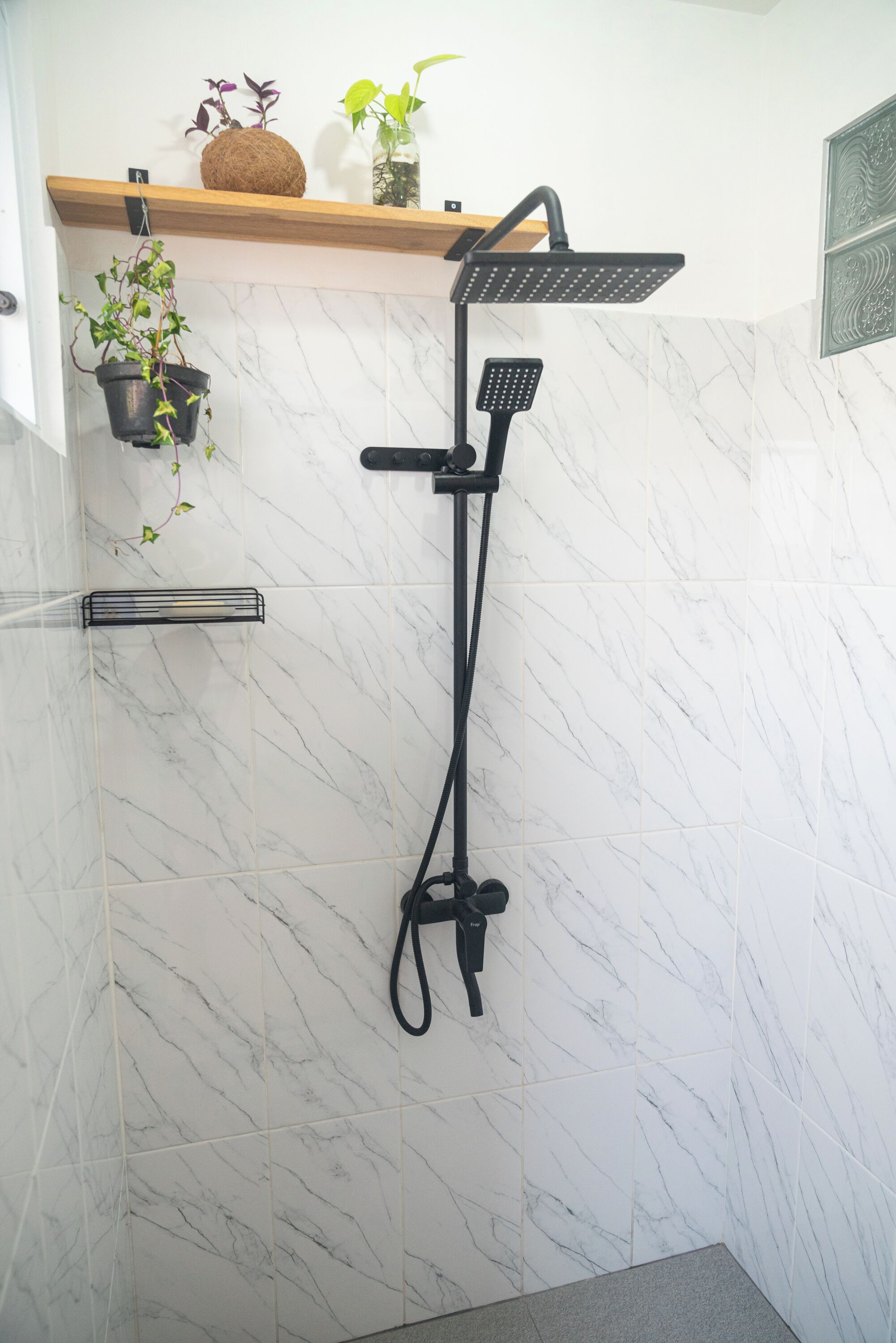 Un baño moderno con un cabezal de ducha negro, una pared de mármol y un estante con plantas.