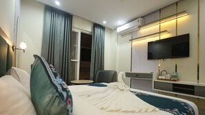 A hotel room with a neatly made bed, a flat-screen TV mounted on the wall, a desk with a chair, and a window with curtains. - Roots & Peaks (Rishikesh)