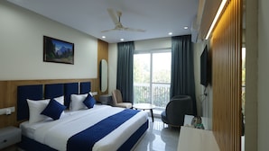 A hotel room with a large bed, a desk, a chair, a TV, and a balcony with curtains. - Roots & Peaks (Rishikesh)