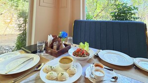 A dining table set with plates, bowls, and utensils, a vase with flowers, and a view of greenery outside. - Roots & Peaks (Rishikesh)