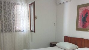 Una habitación de hotel con una cama, una mesita de noche, una ventana con cortinas, un equipo de aire acondicionado y un cuadro en la pared.