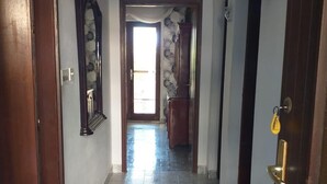 Un pasillo con suelo de baldosas, una puerta de madera a la izquierda, un espejo en la pared y un extintor a la derecha.