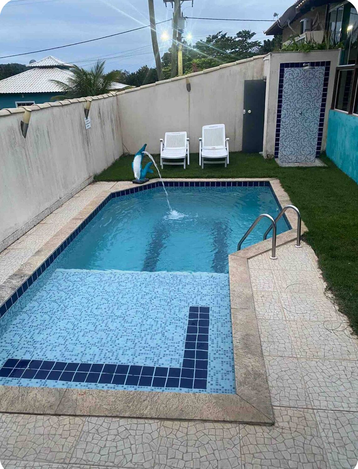 A rectangular swimming pool with a tiled edge, surrounded by a paved area with a mosaic pattern. There are two white chairs and a dolphin-shaped water feature.