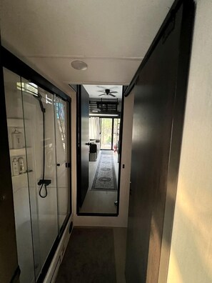 A narrow hallway with a glass shower enclosure, a door leading to a room with a ceiling fan, and a visible outdoor area with a rug. - Luxurious Boutique Tiny House for a Relaxing Getaway in Wimberley, Texas (Wimberley)