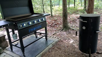 A portable barbecue grill with wheels and a side burner, set in a wooded area.