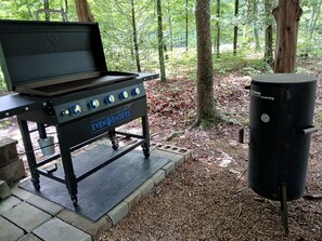 En portabel grill med hjul och en sidobrännare, placerad i ett område med träd och buskar.
