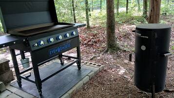 A portable barbecue grill with wheels and a side burner, set in a wooded area.