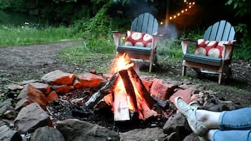 Un feu de camp entouré de rochers et de verdure, avec deux fauteuils Adirondack, dont l’un est doté d’un coussin rouge et l’autre d’un coussin blanc.