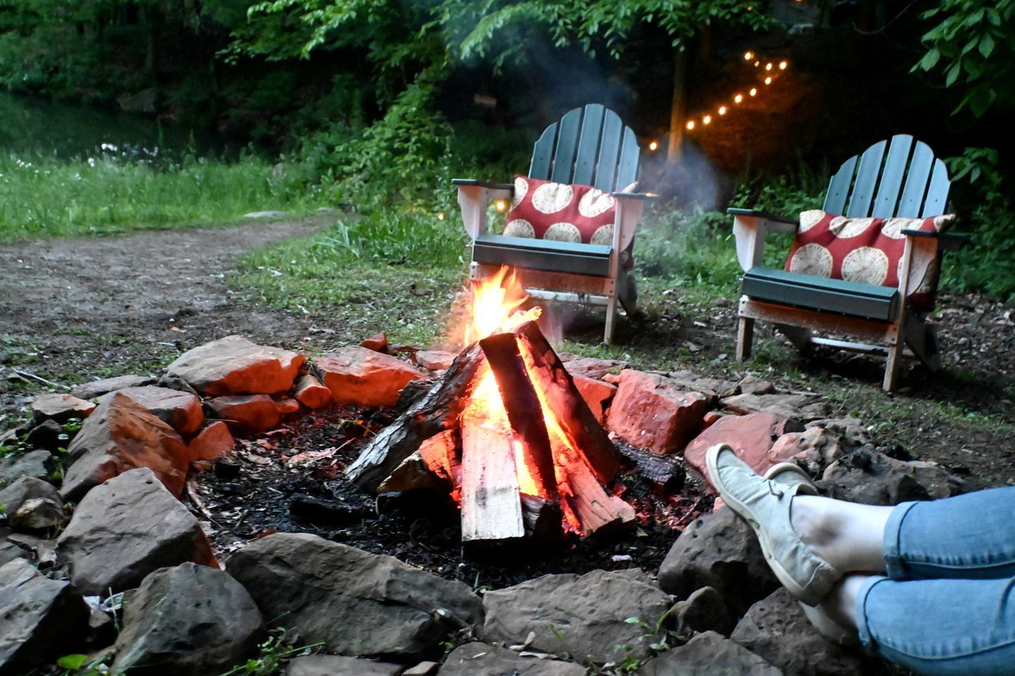 Un feu de camp entouré de rochers et de verdure, avec deux fauteuils Adirondack, dont l’un est doté d’un coussin rouge et l’autre d’un coussin blanc.