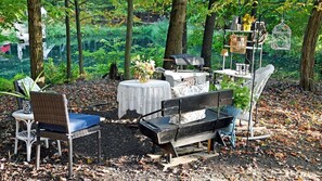 Un espace extérieur aménagé avec des chaises en osier, une table recouverte d’une nappe en dentelle, un barbecue et des objets décoratifs.