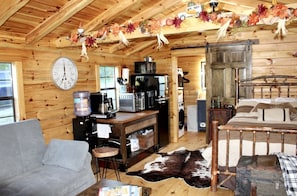 A rustic cabin interior with a living area, kitchen, and a bed with a metal frame.