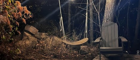 A wooden deck with a swing chair and a bench, surrounded by trees and illuminated by string lights.