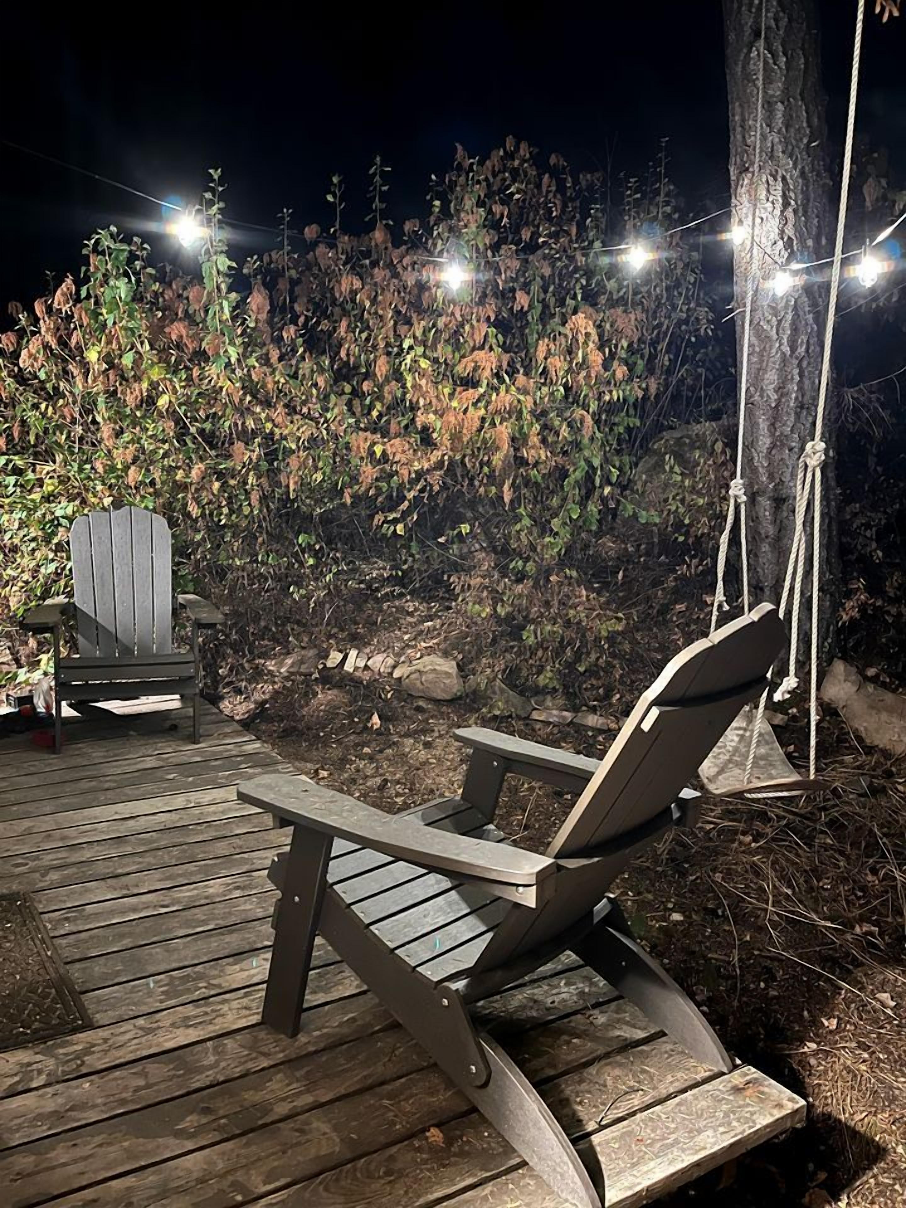 Una terraza de madera con una silla de descanso y una silla Adirondack, rodeada de árboles e iluminada por luces colgantes.