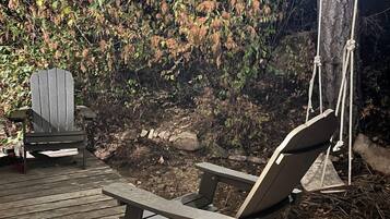 Una terraza de madera con una silla de descanso y una silla Adirondack, rodeada de árboles e iluminada por luces colgantes.