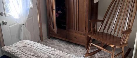 Un dormitorio con una cama, un armario de madera, un sillón mecedora y una pequeña mesa con un letrero.
