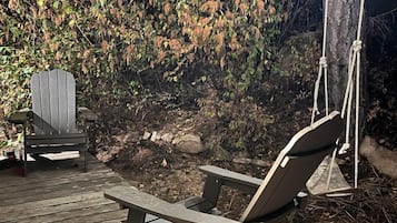 Um deck de madeira com uma espreguiçadeira e uma cadeira Adirondack, cercado por árvores e iluminado por luzes de Natal.