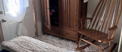 A bedroom with a bed, a wooden wardrobe, a rocking chair, and a small table with a sign.