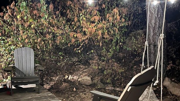 Un patio in legno con una poltrona da relax e una sedia Adirondack, immerso tra gli alberi e illuminato da luci a catena.