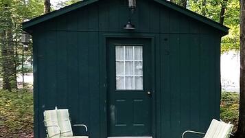 A small green wooden cabin with a door and two white chairs outside.