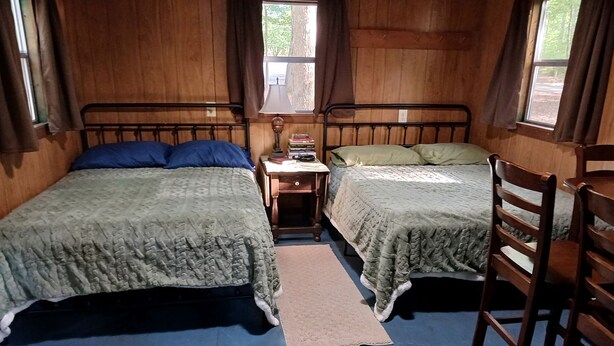 A room with two beds, a small table with books, a chair, and a window.