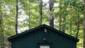 A small green wooden cabin with a door and two white chairs outside.