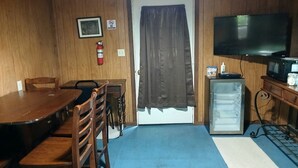 A room with a wooden dining table, chairs, a small refrigerator, a television, and a door with a curtain.