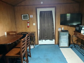 Quarto com mesa de jantar de madeira, cadeiras, uma pequena geladeira, televisão e uma porta com cortina.