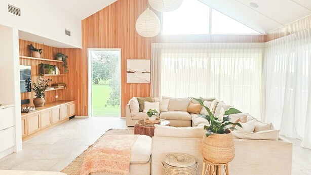 Una sala de estar moderna con un sofá grande, una mesa de centro y un televisor. Se observa un mueble de madera con estantes y una planta. La habitación tiene un ventanal con cortinas.