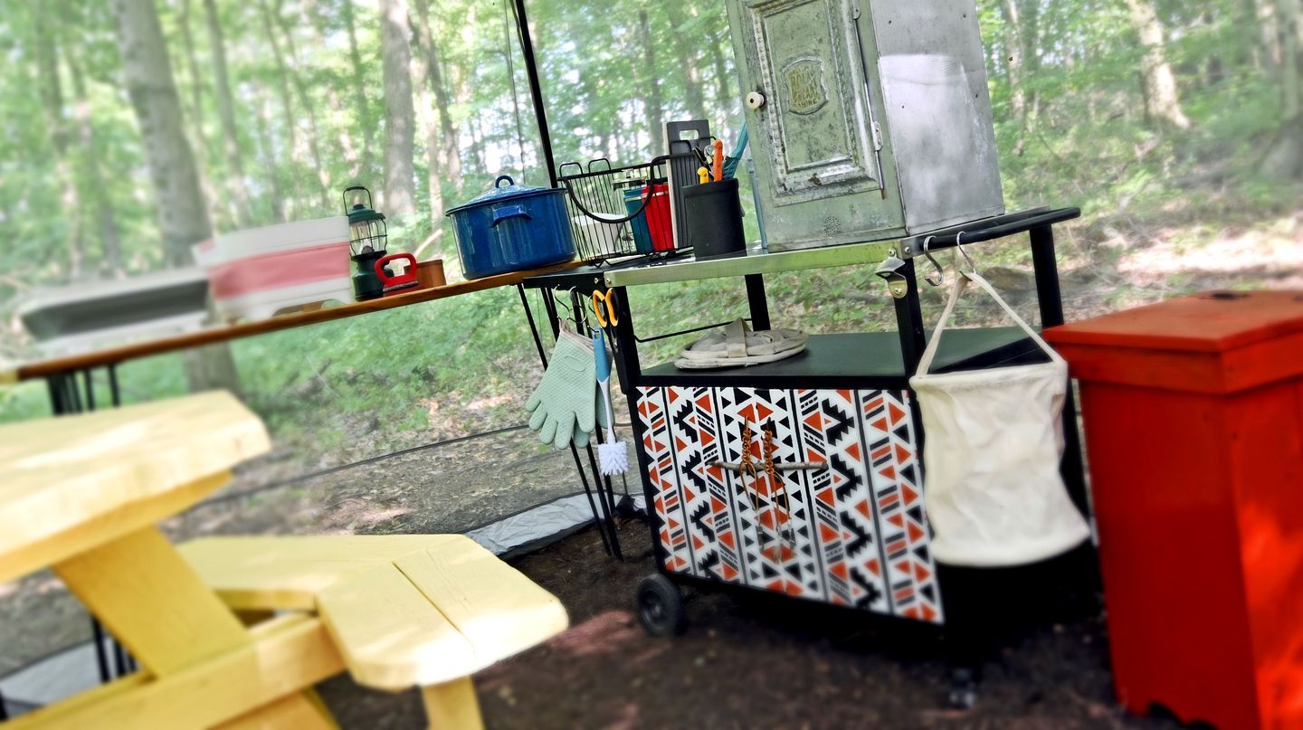 Un campamento con una mesa, un carro con diferentes objetos y un bosque al fondo.