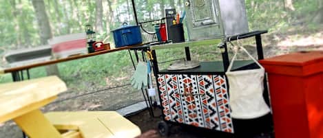 Un campamento con una mesa, un carro con diferentes objetos y un bosque al fondo.