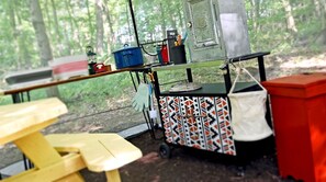 A camping setup with a table, a cart with various items, and a forest background.
