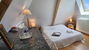 A small, cozy bedroom with a sloped ceiling, a skylight, a bed with neatly folded towels, a bedside table with a lamp, and a radiator. - La Tramolière, accommodation in a green setting (Le Passage)