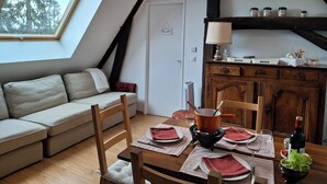 A cozy loft bedroom with a sloped ceiling, a skylight, a wooden dining table set for two, a sofa, and a small kitchen area with a stove and cabinets. - La Tramolière, accommodation in a green setting (Le Passage)