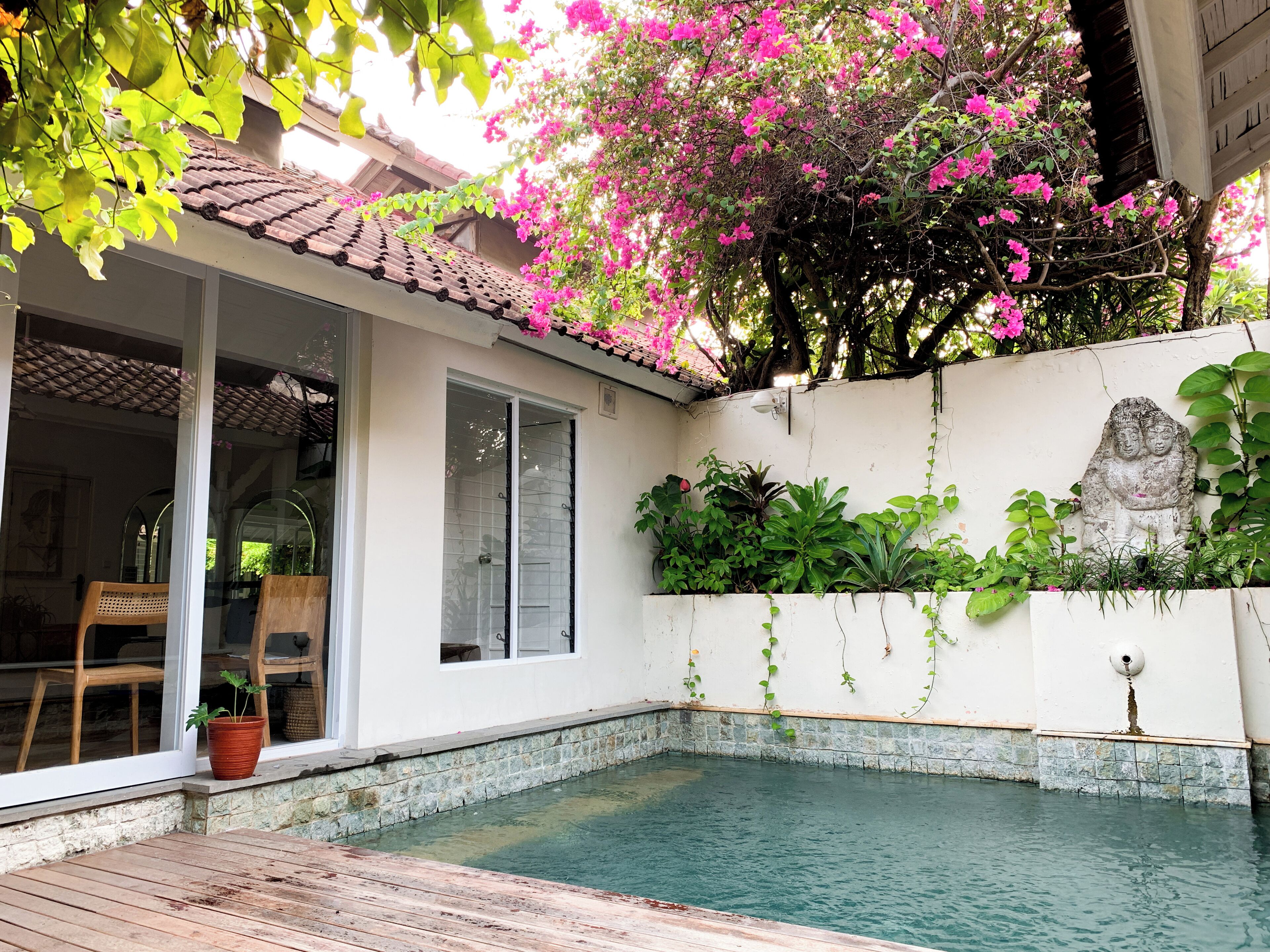 Ein Schwimmbecken mit Fliesen umgeben, eine weiße Wand mit Buddha-Statue und ein Balkon mit Stuhl und Topfpflanze.