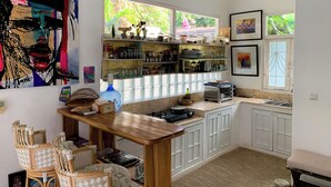 A kitchen with white cabinets, a wooden table, wicker chairs, and a large window with a view of greenery. - Tropical Oasis 2 BR Villa Near Beach (Seminyak)